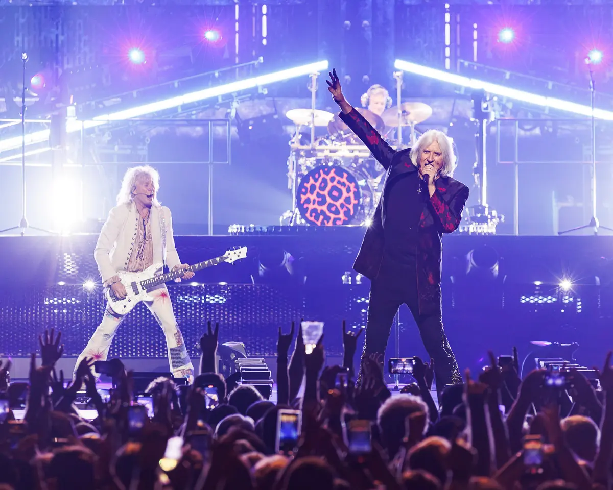 Live rock concert on stage with two performers; singer raises arm while guitarist in white plays a white guitar under purple lights, drummer in back.] , but fix grammar.