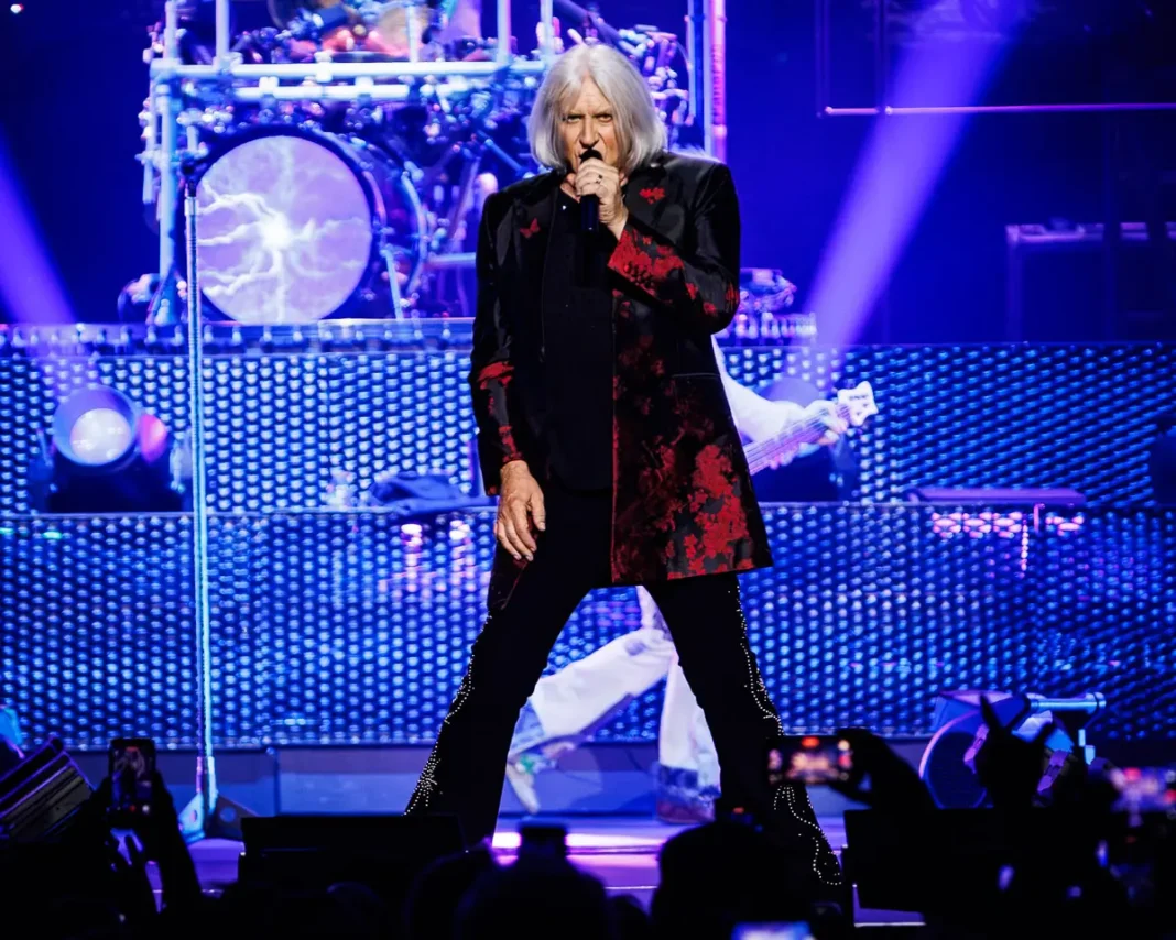 Def Leppard - The O2 Arena - 2 July 2026 Photo Manuela Langotsch 7 Live concert performer with long blond hair, wearing a black and red embroidered jacket, singing into a microphone on a blue-lit stage with drums in the background.