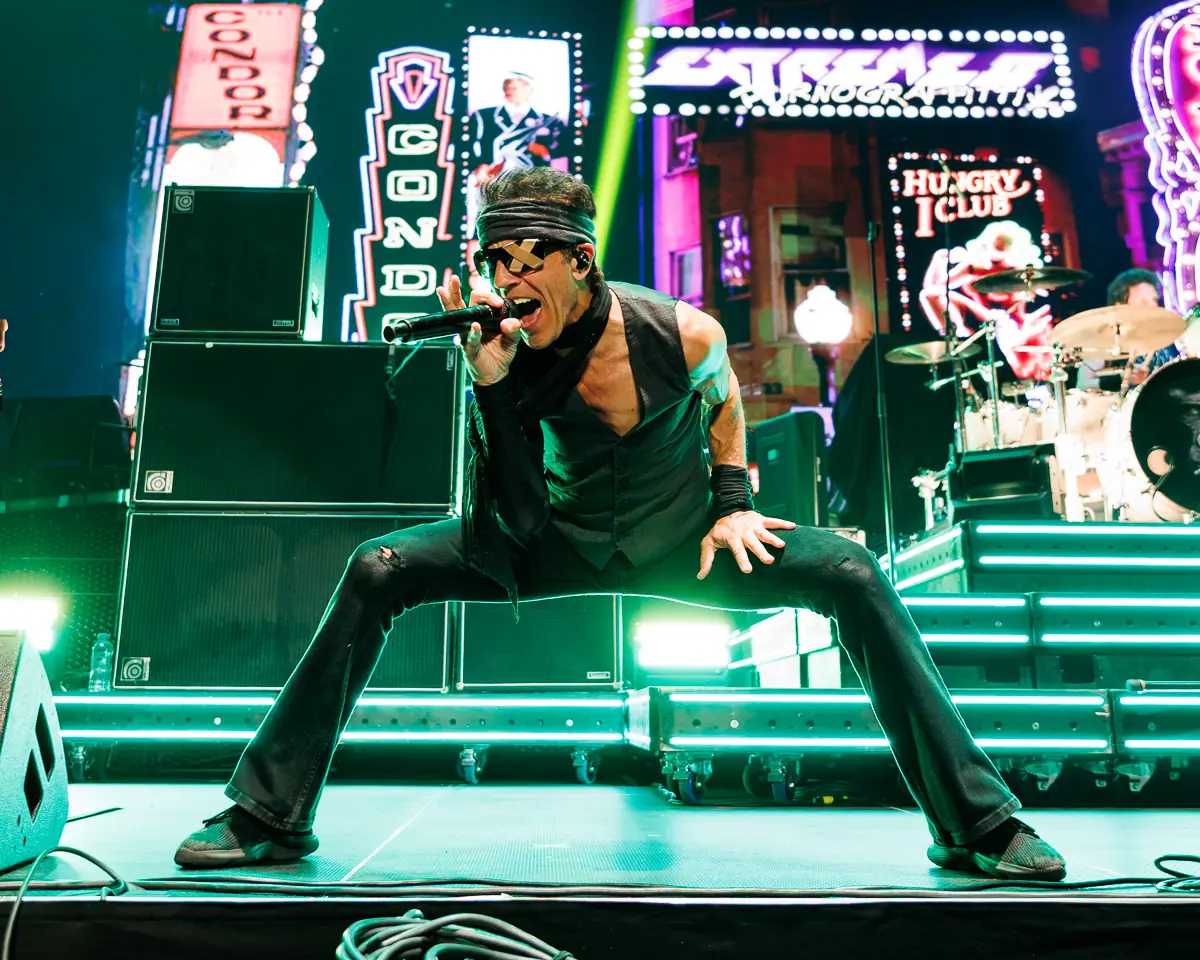 Extreme - The O2 Arena London - 2 July 2026 Photo Manuela Langotsch | MetalTalk - Heavy Metal News Reviews and Interviews Male rock singer crouches center stage, singing into a microphone with neon-lit signs and drums in the background.