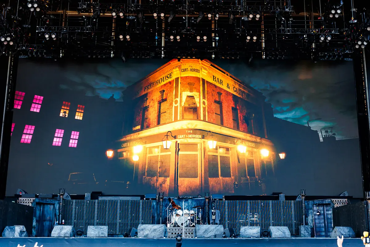Concert stage with a drum kit and a towering screen showing a lit brick pub exterior labeled The Cart & Horses at night