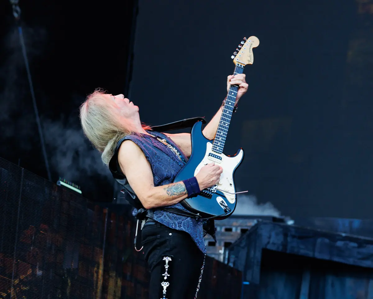 Guitarist with long blond hair wearing a denim vest, playing a blue Stratocaster on stage.
