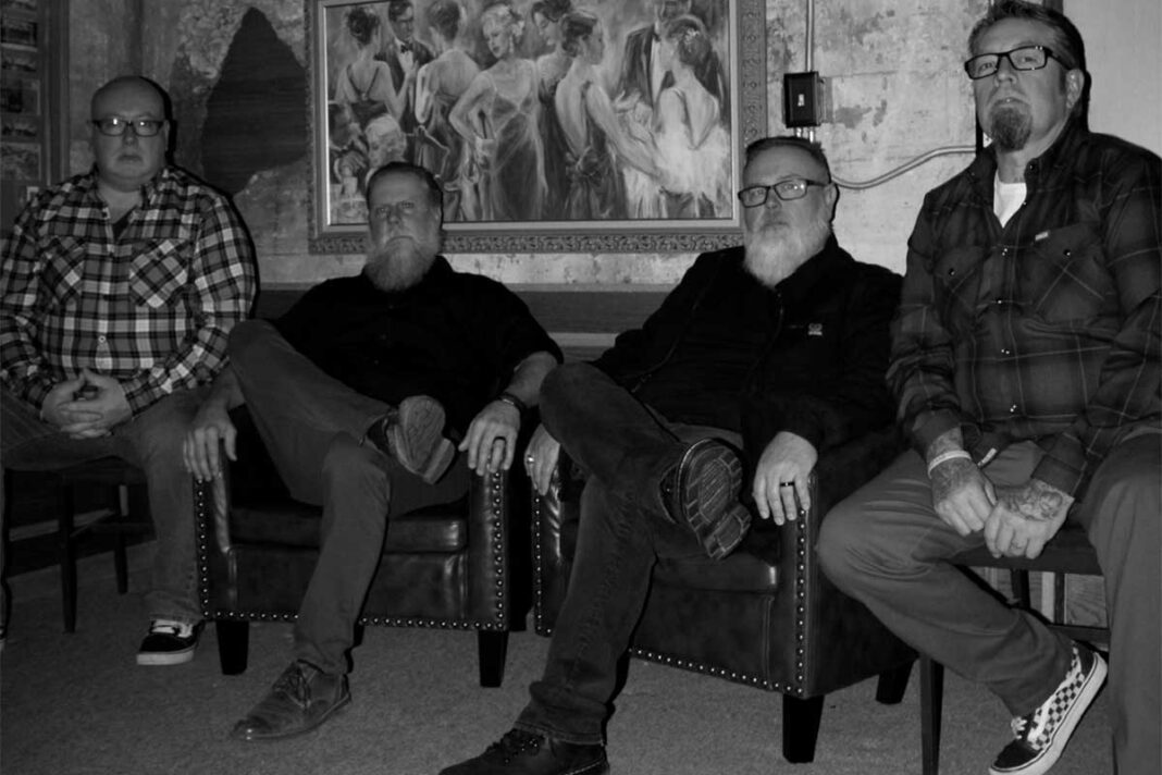 Four men sit on chairs in a dim room, posing for the camera in casual plaid shirts and dark jackets; a large painting is behind them on the wall.